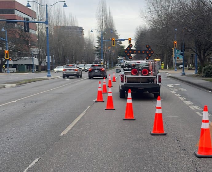 A single-lane closure on a multi-lane urban arterial: an arrow board truck at the head of the closure with a cone taper leading traffic into the adjacent lane, past a marked bike lane.