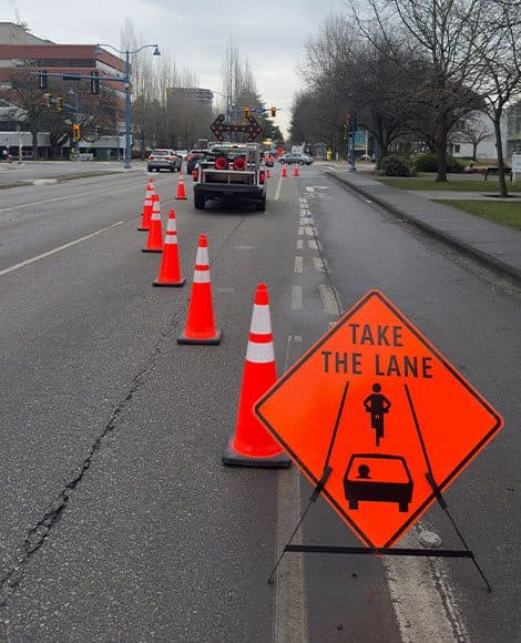A cone taper narrowing an urban arterial to one lane, with an arrow board truck ahead and a TAKE THE LANE sign directing cyclists into the running lane rather than into the closure.