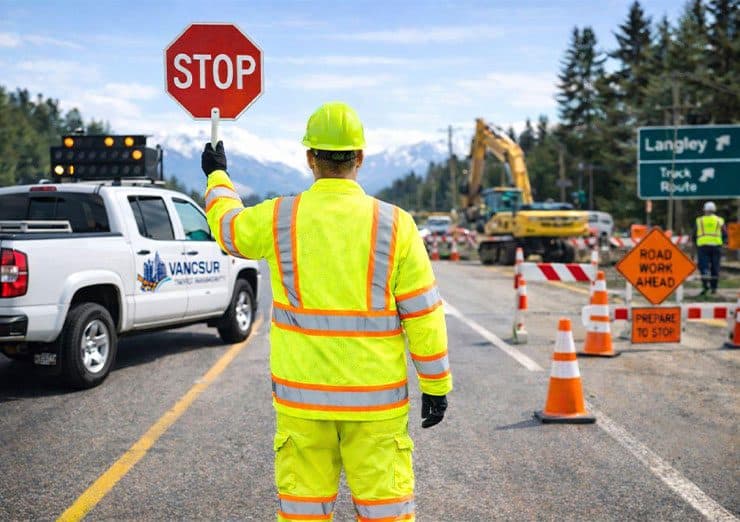 A certified Traffic Control Person in high-visibility PPE holding a STOP paddle on a two-lane highway, with an arrow board, cones and an excavator working behind the taper.