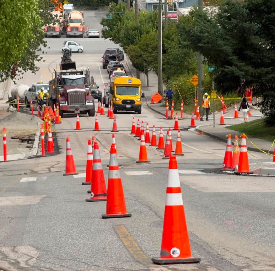 A long single-lane closure on a residential arterial: two rows of cones taper traffic into one lane past excavation work, with a dump truck, a concrete mixer and crew in high-visibility apparel working behind the barrier.